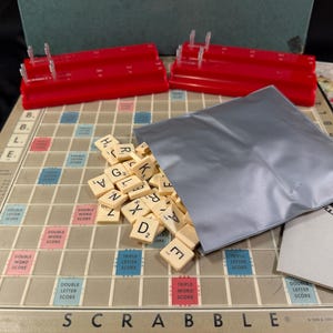 May include: A Scrabble board game set with a beige game board, red tile racks, and a gray pouch filled with letter tiles. The game board has colored squares for bonus points. A Scrabble guide is visible.