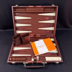 May include: Open brown leather backgammon set with white and brown game pieces, dice, and a rule book. The set includes two brown leather dice cups and is displayed on a black surface.