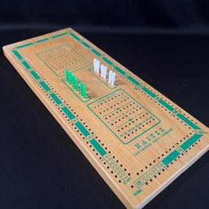 May include: A wooden cribbage board with a green track and white and green pegs. The board has a continuous track and the text "MASTER CONTINUOUS TRACK CRIBBAGE" printed on it.