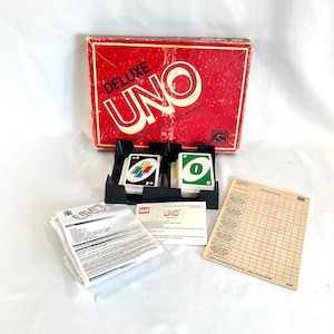 May include: A vintage UNO Deluxe game box with a red lid and the word "DELUXE UNO" printed in white. The box is open and the game cards are visible. The cards are in a black plastic tray with two compartments. The game includes a scorecard and instructions.