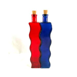 May include: Two decorative glass bottles, one red and one blue, with a wavy design. Each bottle has a cork stopper. The bottles are side-by-side against a white background.