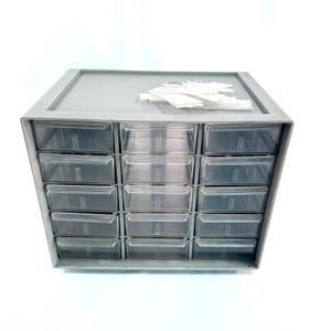May include: A gray plastic parts organizer with sixteen clear plastic drawers. The organizer has a flat top and a gray frame. A small pile of white labels rests on top of the organizer.