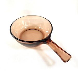 May include: A vintage amber-colored glass saucepan with a handle. The pan is round with a slightly flared rim and a long, integrated handle. The glass has a warm, translucent hue, and the pan is set against a white background.