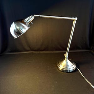 May include: A silver-toned desk lamp with an adjustable arm and a rounded shade. The lamp has a circular base and a cord extending from the base. The lamp is made of metal and has a polished finish.