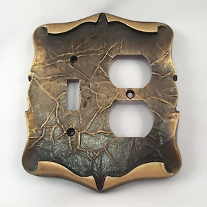 May include: A decorative brass outlet cover with a textured, hammered finish. The cover has a single light switch and a double outlet.