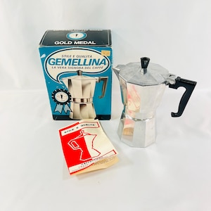 May include: A silver Gemellina stovetop espresso maker with a black handle and a black knob. The coffee maker is next to a blue and gold box with the words "Gold Medal" and "Gemellina" printed on it. A red and white paper with the words "Stile e Qualita" is also in the image.