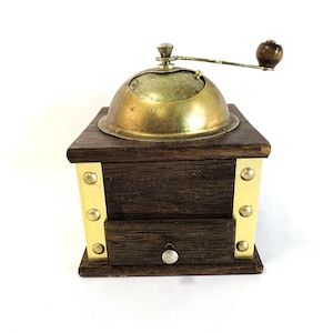 Puede incluir: Un molinillo de café antiguo con una base de madera oscura, detalles de latón y una parte superior en forma de cúpula de latón. El molinillo cuenta con una manivela con un mango de madera y un pequeño cajón. El diseño es clásico y funcional.
