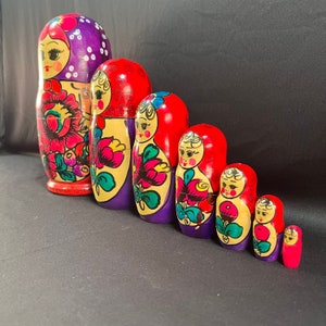 May include: A set of eight traditional Russian nesting dolls, also known as matryoshka dolls. The dolls are painted in vibrant colors with floral patterns and feature the faces of women. The largest doll is purple with red accents and the smallest doll is red with white accents.