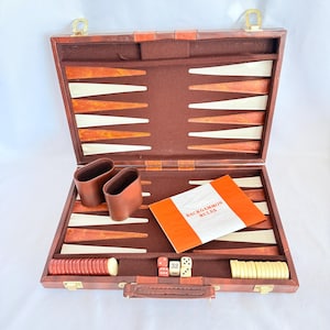 May include: Open backgammon set in a brown leather case. The set includes brown leather dice cups, dice, and game pieces in red, white, and cream colors. A booklet with the text "BACKGAMMON RULES" is also included.