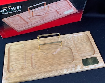 May include: A wooden valet tray with two gold metal handles. The tray has four compartments, including a small rectangular compartment with a gold metal nameplate. The tray is made of solid oak.