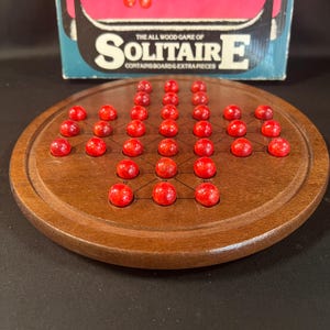 May include: A vintage wooden Solitaire game with a round, dark brown board and red marbles. The game board has a series of drilled holes for the marbles. The box in the background reads "The All Wood Game of Solitaire E".