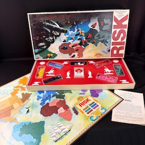 May include: Vintage Parker Brothers Risk World Conquest Game. The game box is open, revealing a red interior with game pieces in clear containers. The game board features a world map with continents and territories. The box lid displays a cannon and the word "RISK".