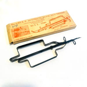 May include: A black metal hook rug needle with a metal frame and a needle. The needle is attached to the frame with a screw. The needle is sharp and pointed. The frame is rectangular with rounded corners. The needle is used to make rugs. The box reads "No. 3-SO. E. Z. AUTOMATIC HOOK RUG NEEDLE 25¢ THIS INSTRUMENT IS ESPECIALLY ADAPTED FOR MAKING HOOK RUGS OF SO. E. Z. YARN For longer stitch use No. 7 Needle WONDERART 2212 Byron St. Chicago, Ill."