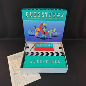 May include: A Guesstures board game set featuring a teal box with a blue gradient lid displaying stylized figures. Inside is a teal plastic action timer shaped like a movie clapperboard, a red card box, and a white instruction manual with black text.