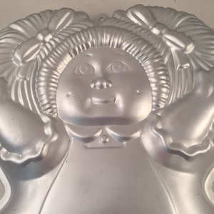 May include: A silver metal cake pan shaped like a girl with pigtails and a bow. The pan has a detailed face with a smile and rosy cheeks.