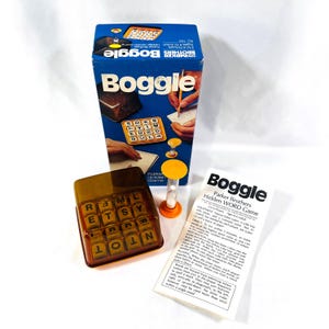 May include: A vintage Boggle game by Parker Brothers. The game includes a clear plastic cube tray with letter tiles, a sand timer, and instructions. The box is blue and white with the game's name in bold, white letters.