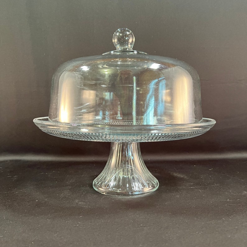 May include: A clear glass cake stand with a dome lid. The stand has a fluted base and a wide, flat top.