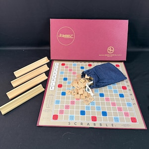 May include: A vintage Scrabble board game set. The set includes a maroon box, a game board with red and blue squares, wooden letter tiles, and wooden tile racks. The game board has the word "SCRABBLE" printed on it.