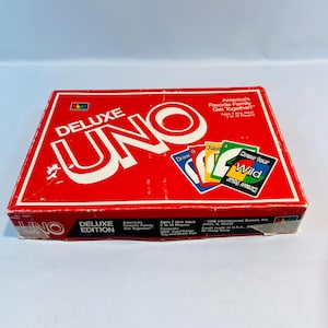 May include: A red box for the Deluxe Edition of the card game UNO. The box features the UNO logo in white and the text "America's Favorite Family Get Together!" in white. The box also shows four UNO cards: a Draw 8 card, a Reverse card, a Draw Four card, and a Wild card.