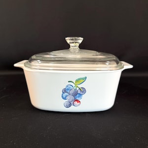 May include: White casserole dish with a clear glass lid. The dish features a painted bunch of blue grapes with green leaves and a red berry. The dish has two handles and a clear glass knob on the lid.