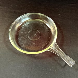 May include: Clear glass skillet with a textured bottom and a handle. The skillet has a round shape and a handle with vertical ridges. The word "Rangecraft" is embossed in the center of the skillet.