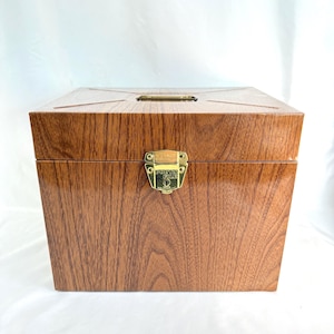 May include: A wooden box with a glossy finish and a wood grain pattern. The box has a gold-colored latch and a small gold-colored metal plate on the top. The box is rectangular and has a hinged lid.