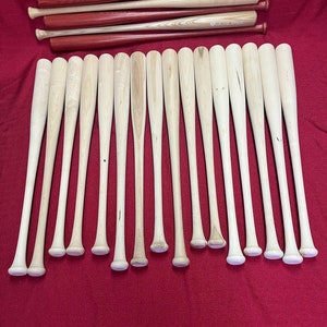 May include: A row of 16 unfinished wooden baseball bats, all lined up on a red surface. The bats are all the same size and shape, and they are all made of a light-colored wood. The bats are all in focus, and the background is blurred.