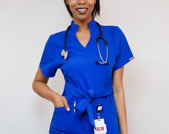May include: A woman wearing a royal blue medical scrub top and matching pants. The top has a V-neck and a white tie at the waist. She is wearing a stethoscope around her neck and a name tag that says "RN".