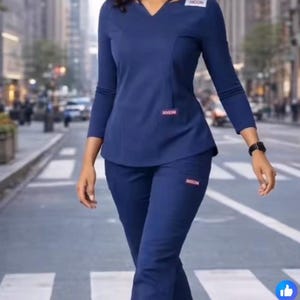 May include: Navy blue medical scrubs set. The outfit includes a long-sleeved top with a V-neck and matching pants. The scrubs have the brand name "Jocccini" on the top and pants. The person is wearing a black watch and gray sneakers.