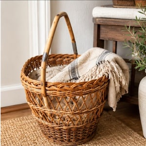 May include: A round, woven brown wicker basket with a single arched handle. The basket features an open-weave pattern and contains a folded, textured beige throw blanket with a dark grey stripe and fringed edges draped over the side.