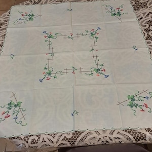 May include: A square, cream-coloured tablecloth with embroidered floral designs in green, red, and blue. The embroidery features a central square border and corner accents. The edges are finished with a green scalloped trim.