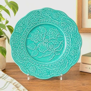 May include: A round, turquoise ceramic plate featuring a raised, embossed floral and leaf pattern in the centre, surrounded by a decorative border of intricate lattice and petal designs with a scalloped edge, displayed on a clear stand.