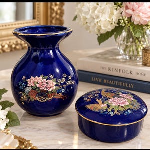 May include: A deep blue ceramic vase and a matching round lidded trinket box. Both pieces feature gold-trimmed edges and intricate, colourful floral patterns with pink peonies and small blossoms. The vase has a wide, flared rim and a rounded body.
