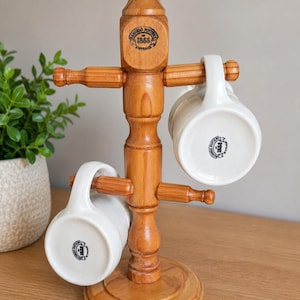 May include: A vertical wooden mug tree with a round base and four horizontal pegs. Two white ceramic mugs with the text Bendigo Pottery 1858 Australia printed on the bottom are hanging from the pegs. The wood has a polished brown finish.