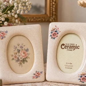 May include: Two rectangular ceramic picture frames with a cream-coloured crackle glaze finish. Each frame features an oval opening and floral decals in the corners. The right frame displays text reading Kiln Fired Ceramic Frame 3 1/2 x 5 in.