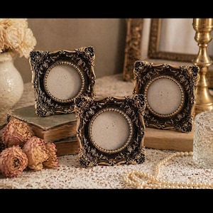 May include: Three decorative square picture frames with a dark bronze finish and floral detailing. Each frame has a circular aperture for a photo. The frames are arranged on a table with vintage decor.