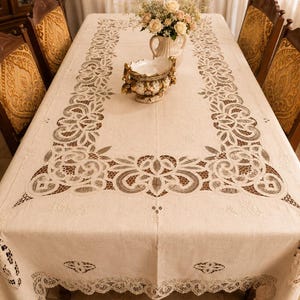 May include: A rectangular off-white tablecloth featuring intricate cutwork embroidery and lace detailing along the borders and centre panel. The fabric displays a woven texture with floral motifs and openwork patterns, draping over a dining table.