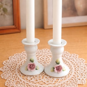 May include: Two white ceramic candle holders with pink rose and green leaf details. Each holder supports a tall, white candle. The candle holders rest on a beige lace doily, creating a vintage aesthetic.