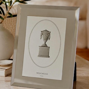 May include: A rectangular, grey picture frame with a mirrored finish. It contains a black and white print of a classical urn on a pedestal, within an oval dotted border. The word "WEDGWOOD" is printed below the image.