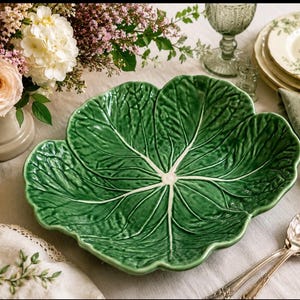 May include: A glossy green ceramic serving bowl shaped like a large cabbage leaf. The bowl features a realistic embossed texture with prominent white veins radiating from the centre, mimicking the natural appearance of a leafy vegetable.