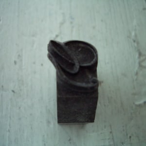 May include: A dark, rectangular metal stamp with a decorative, curved design on top. The stamp is likely used for imprinting or embossing. The object is a dark, muted color, possibly iron or steel.