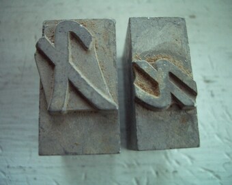 May include: Two weathered, rectangular metal printing blocks. One block features a stylized letter "K", the other a letter "S". The blocks are a dull gray color, showing signs of age and use, with some surface imperfections.