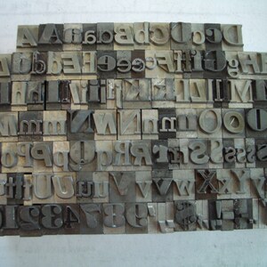 May include: A collection of vintage metal letterpress printing blocks, arranged in a grid. The blocks are various sizes and feature uppercase and lowercase letters, numbers, and symbols. The metal has a worn, aged appearance.