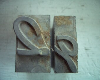 May include: Two weathered, gray metal letterpress printing blocks. One block features a stylized letter "Q", and the other a letter "R". The blocks have a rectangular shape and show signs of age and use.