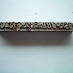 May include: A metal type set with uppercase letters arranged in a row. The letters are in a serif font and appear to be vintage. The metal has a worn, aged appearance.