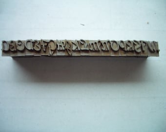 Può includere: Un set di caratteri in metallo con lettere maiuscole in un carattere serif. Le lettere sono disposte in fila e presentano un aspetto usurato e invecchiato.