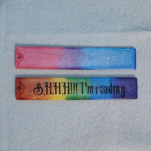 May include: Two rainbow glitter bookmarks. The top bookmark is pink, purple, blue and has a silver glitter finish. The bottom bookmark is a rainbow of colours with the text "SHHH!!! I'm reading." in black.