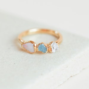 May include: A gold ring with three different colored gemstones. The ring features a pink, blue, and clear stone.