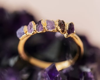 Può includere: Un anello d'oro con una fila di pietre preziose viola e marroni. L'anello è posto su un letto di cristalli viola.