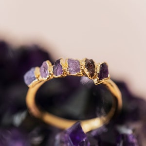May include: A gold ring with a row of purple and brown gemstones. The ring is set on a bed of purple crystals.